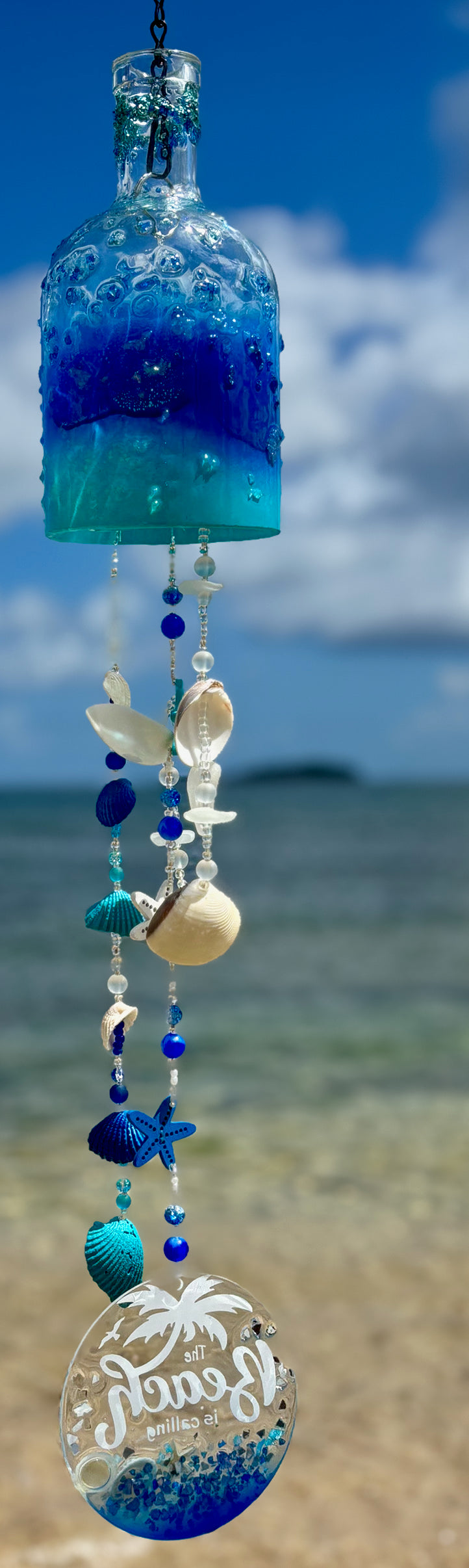 Decorative wind chime with blue and clear design, seashells, and 'Love' charm against a beach background.