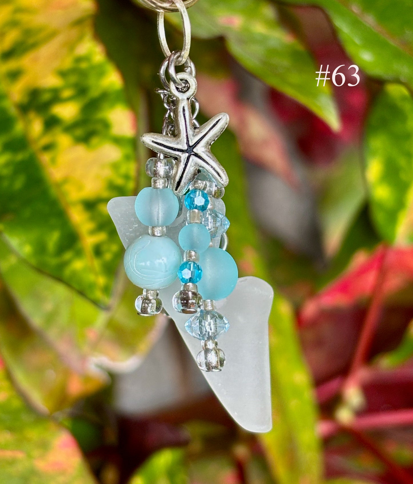 Decorative keychain with blue beads, starfish charm, and white leaf-shaped pendant on a green leaf background.