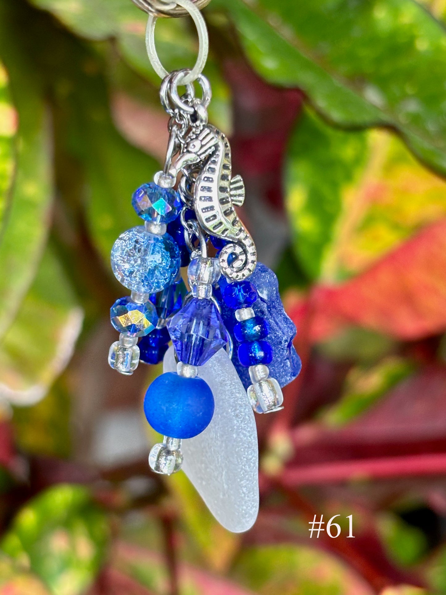 Decorative keychain with blue beads, a seashell, and a seahorse charm against a leafy background.