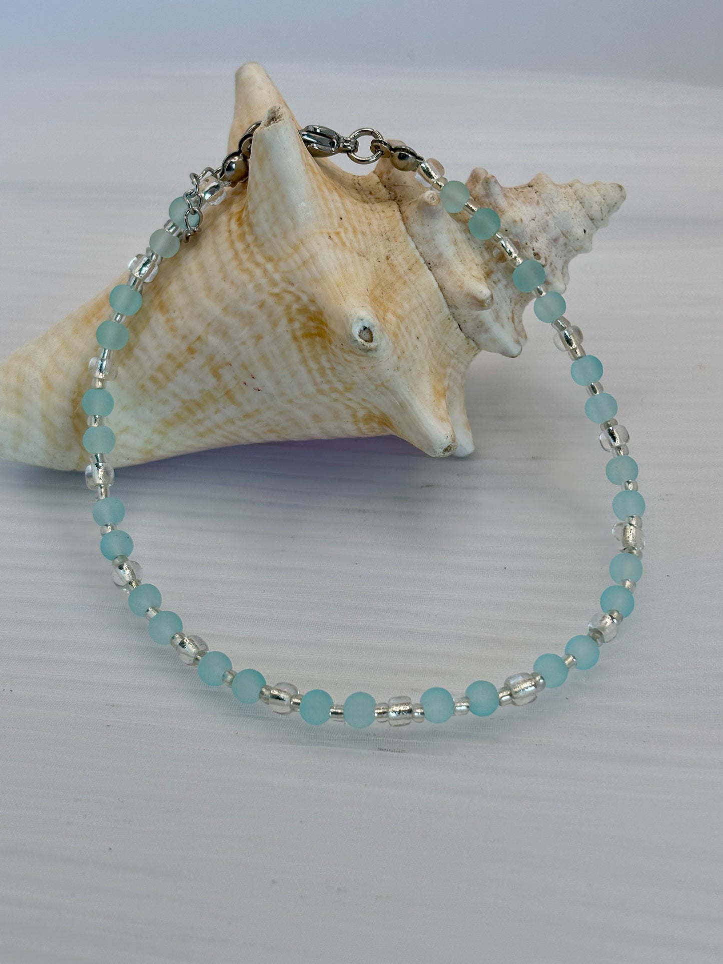 Turquoise beaded anklet on a white surface with a large shell in the background
