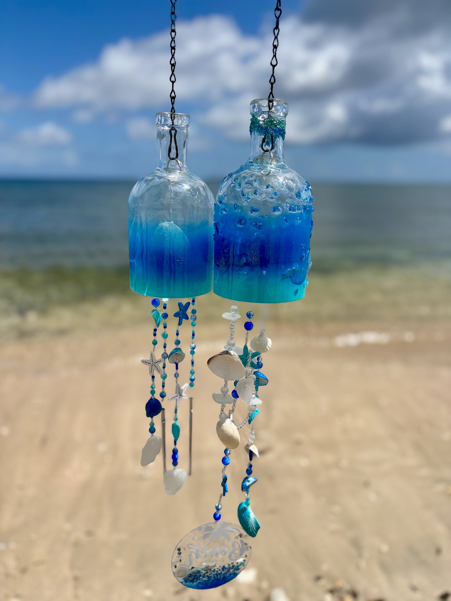 Two rum bottle wind chimes with decorative elements hanging against a beach background.
