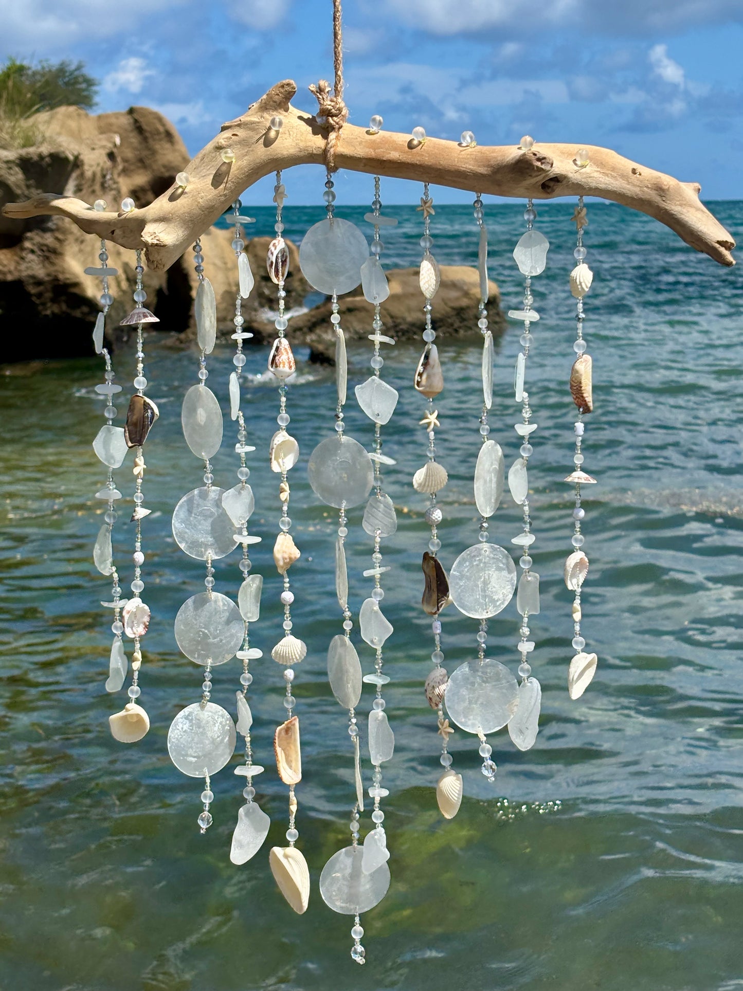 Decorative wind chime with seashells and beads hanging by a body of water.