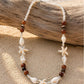 A beaded necklace with starfish and shell charms, displayed on a piece of driftwood over a sandy beach with the ocean in the background.