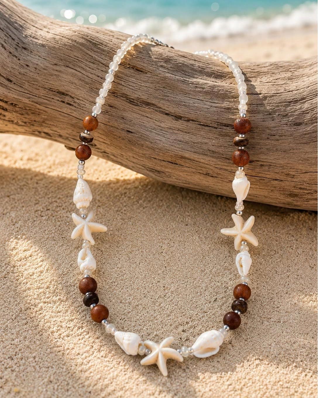 A beaded necklace with starfish and shell charms, displayed on a piece of driftwood over a sandy beach with the ocean in the background.