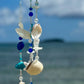 Decorative wind chime with blue and clear design, seashells, and 'Love' charm against a beach background.