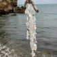 A hanging beach wind chime made from seashells and driftwood, suspended over a rocky shoreline and ocean background.
