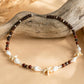 A beaded necklace with dark brown and reddish-brown round beads, white shell pieces, and starfish charms, displayed on a beige fabric surface with a piece of driftwood.