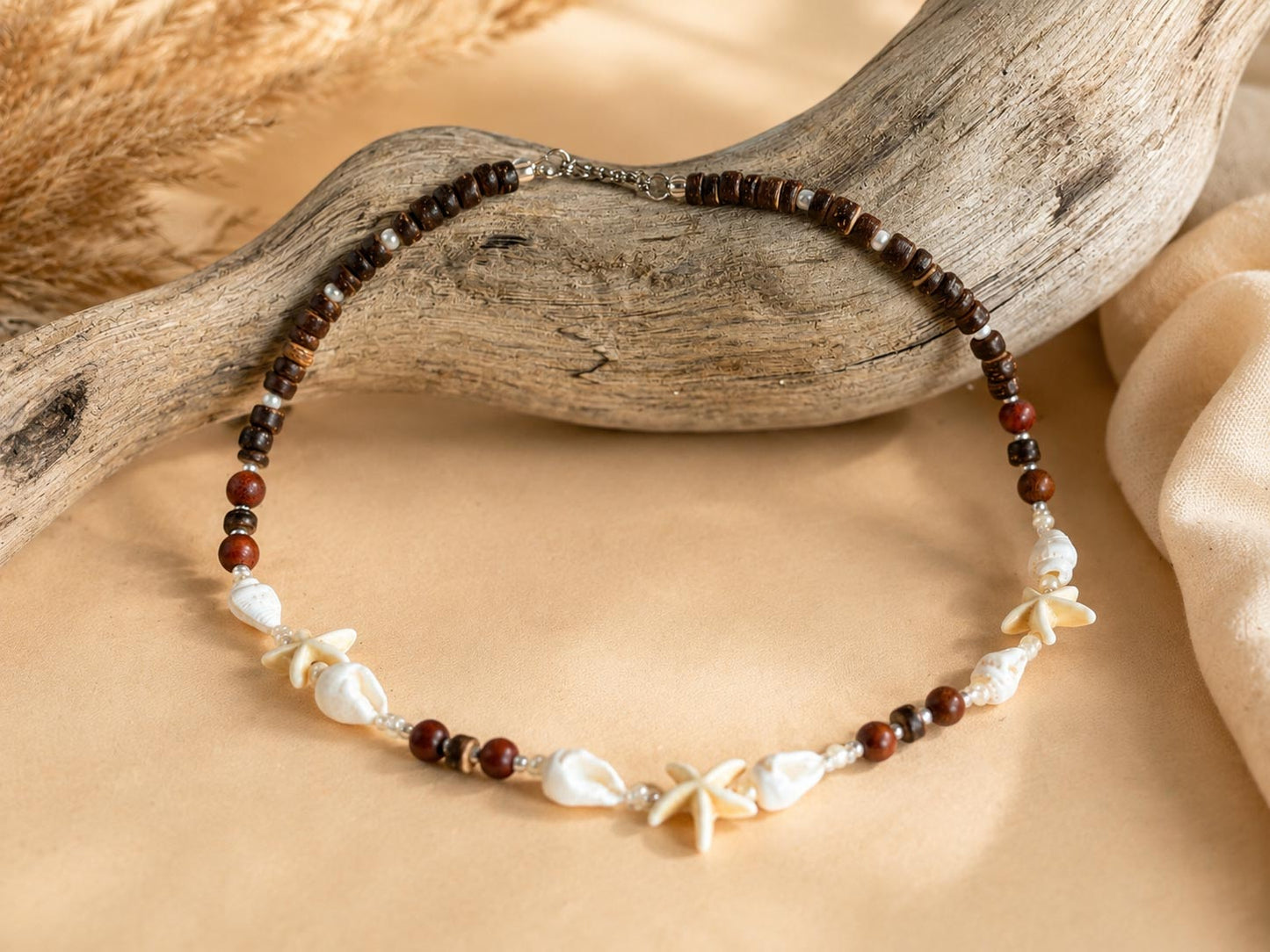 A beaded necklace with dark brown and reddish-brown round beads, white shell pieces, and starfish charms, displayed on a beige fabric surface with a piece of driftwood.