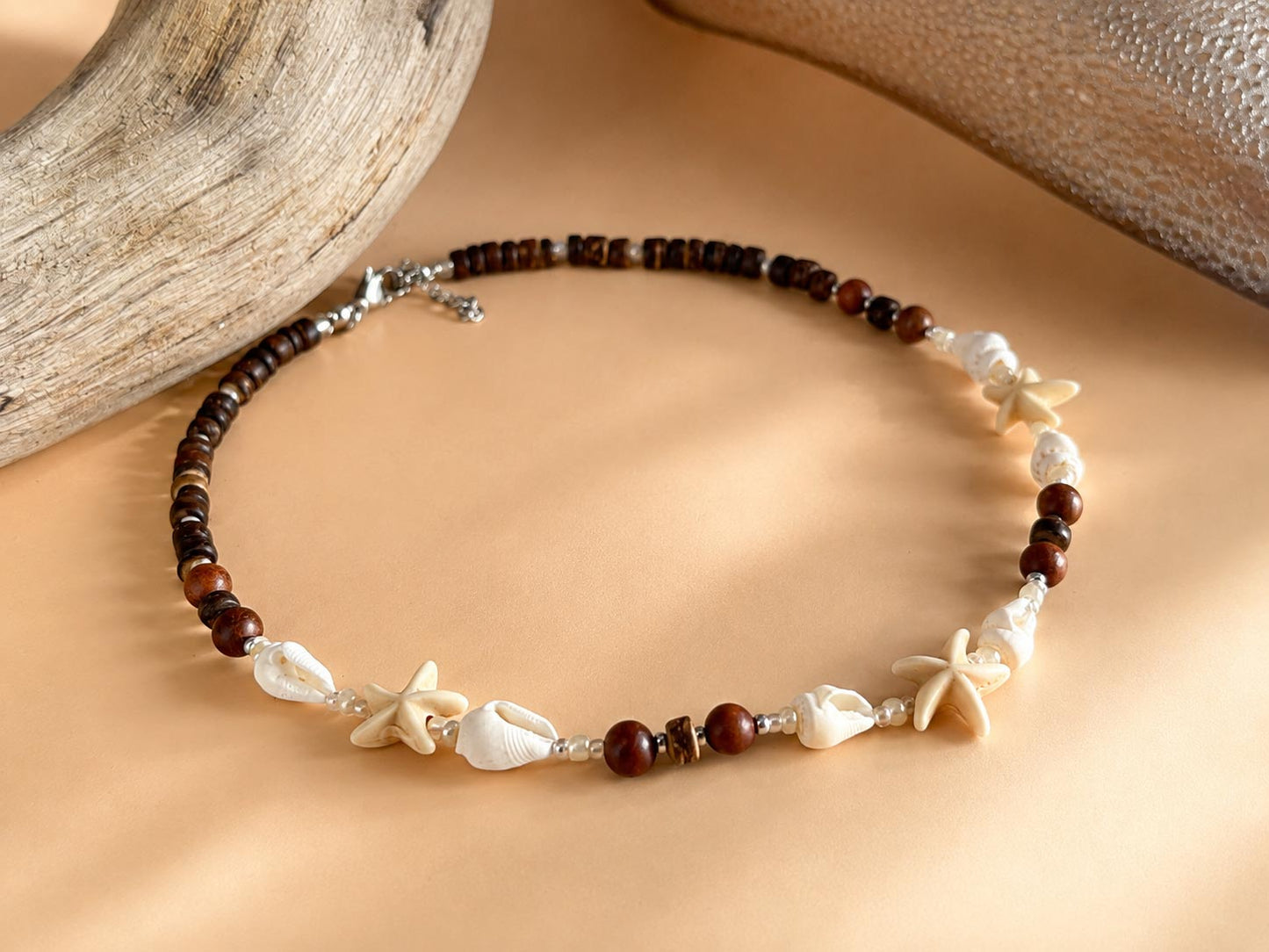 A beaded bracelet with dark brown wooden beads, white seashells, and starfish charms, displayed on a beige background with driftwood props.