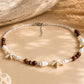A bohemian-style beaded necklace featuring brown wooden beads, white seashells, and white starfish charms, displayed on a beige surface with rustic wood and dried grass props.