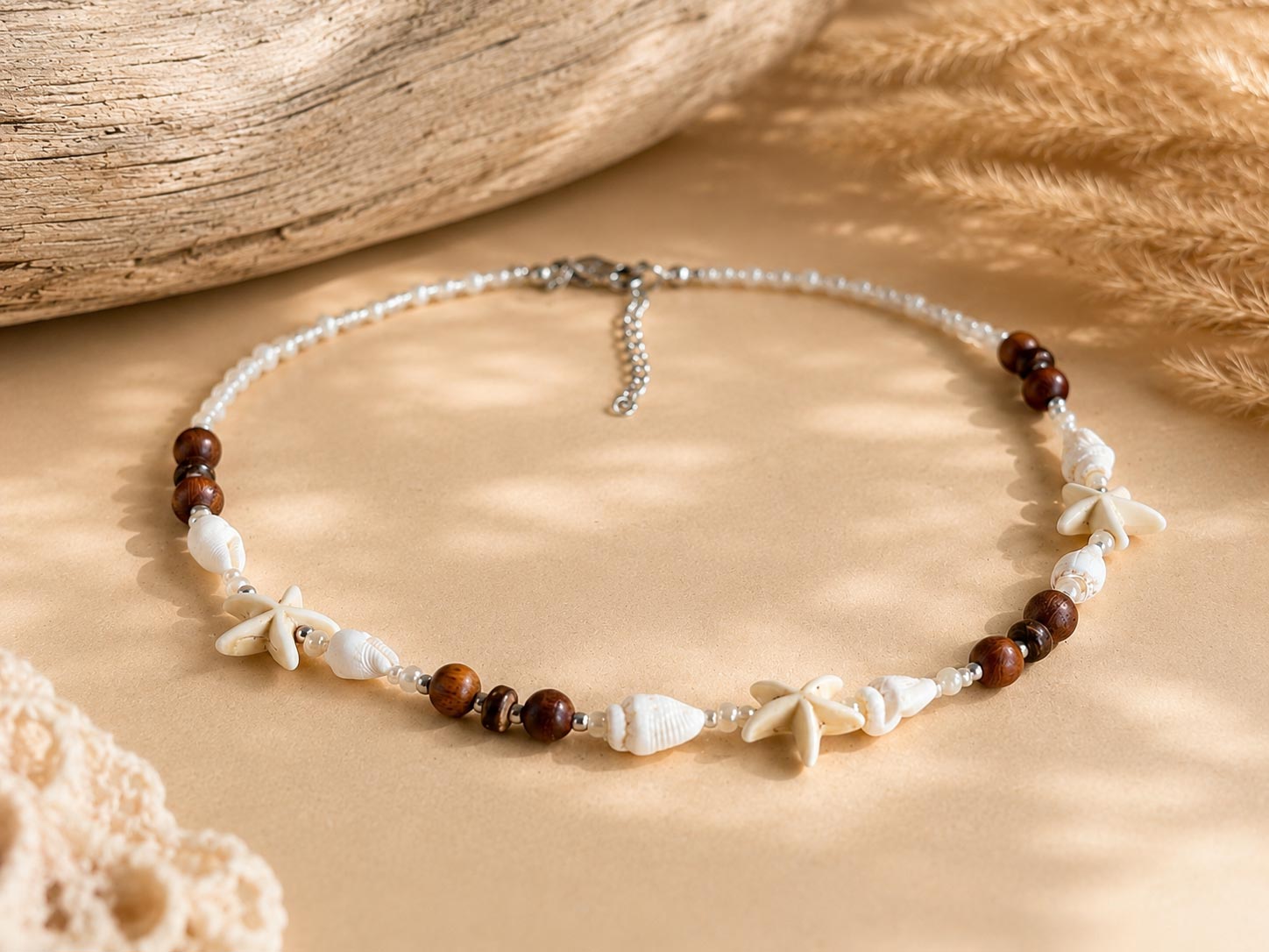 A bohemian-style beaded necklace featuring brown wooden beads, white seashells, and white starfish charms, displayed on a beige surface with rustic wood and dried grass props.