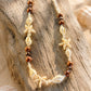 A bohemian-style beaded necklace featuring brown wooden beads, white shell beads, and natural starfish and seashell charms, displayed on a piece of weathered driftwood.