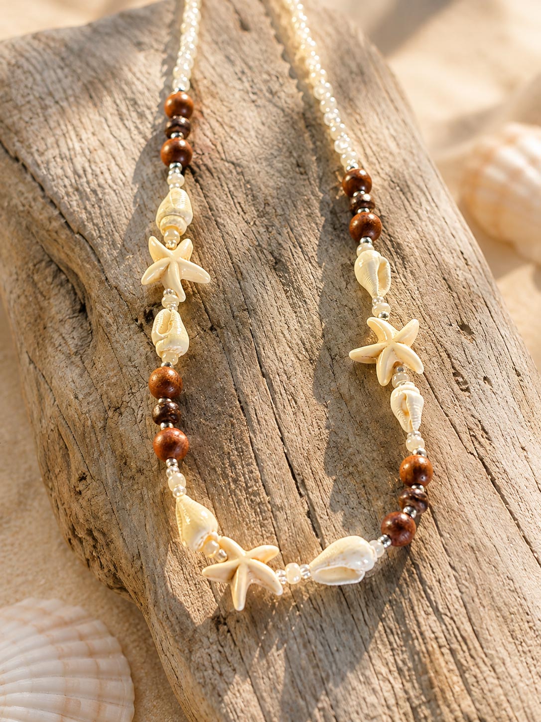 A bohemian-style beaded necklace featuring brown wooden beads, white shell beads, and natural starfish and seashell charms, displayed on a piece of weathered driftwood.