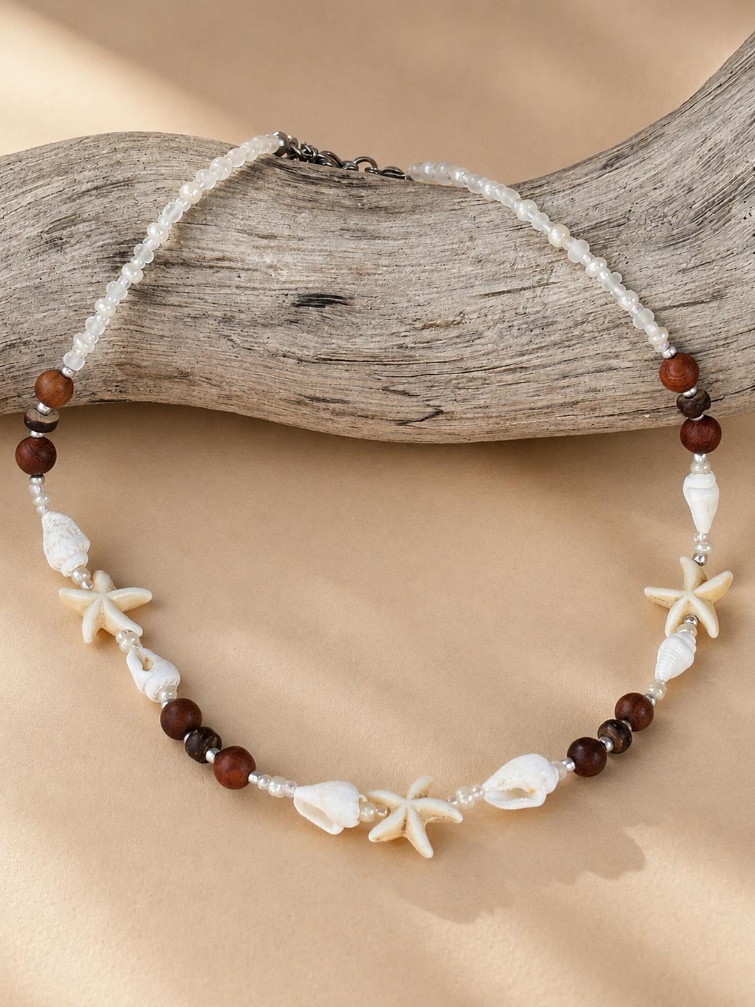 A beaded necklace with white seed beads, dark brown round beads, white seashells, and starfish charms, displayed on a piece of driftwood over a beige fabric background.