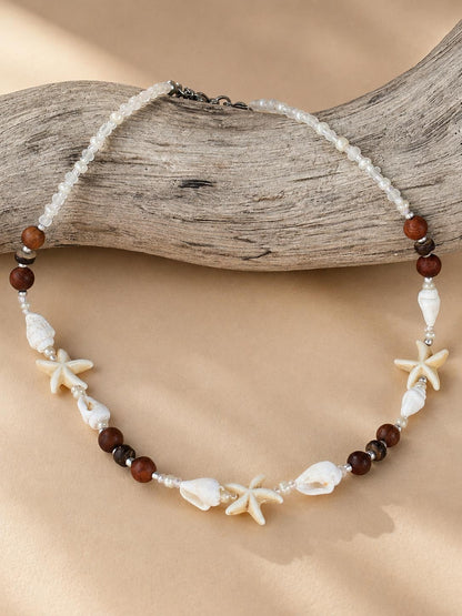 A beaded necklace with white seed beads, dark brown round beads, white seashells, and starfish charms, displayed on a piece of driftwood over a beige fabric background.