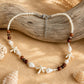 A beaded necklace with white shell beads, brown wooden beads, and starfish charms, displayed on a piece of driftwood over a sandy background with seashells.