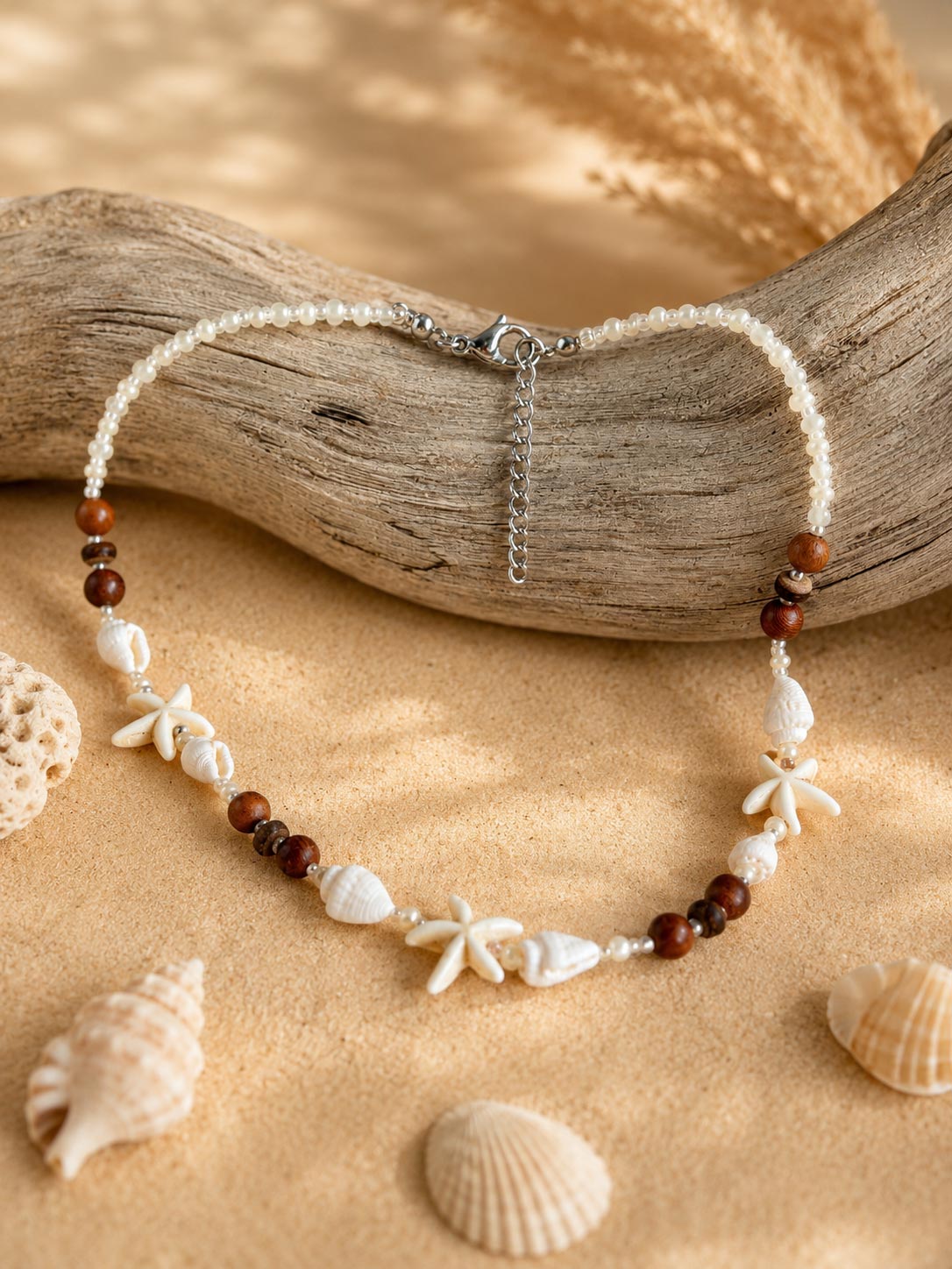 A beaded necklace with white shell beads, brown wooden beads, and starfish charms, displayed on a piece of driftwood over a sandy background with seashells.
