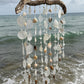 A driftwood beach wind chime with hanging seashells and clear glass beads, suspended over the ocean waves.