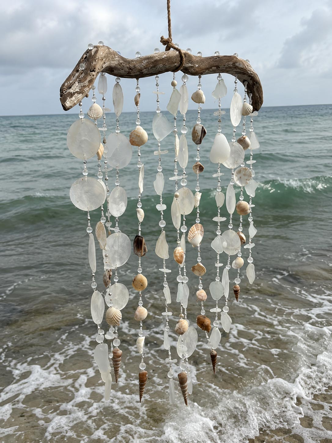A driftwood beach wind chime with hanging seashells and clear glass beads, suspended over the ocean waves.