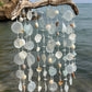 A beach-themed wind chime hanging from a natural driftwood branch, featuring glass discs, seashells, and small beads, set against a blurred ocean background.