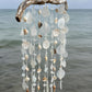 A hanging beach-themed wind chime made from driftwood, translucent glass discs, and natural seashells, suspended against an ocean background.