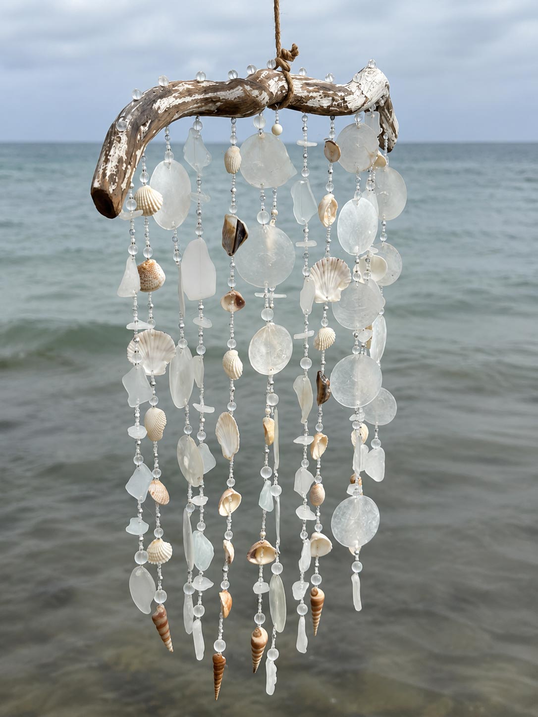 A hanging beach-themed wind chime made from driftwood, translucent glass discs, and natural seashells, suspended against an ocean background.