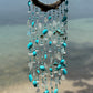 Decorative wind chime with turquoise and white beads and shells against a blurred beach background.